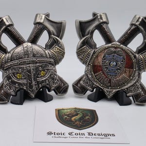 May include: Two metal challenge coins featuring Viking helmet and shield designs, each with crossed axes. The helmet coin has yellow eyes, while the shield coin displays a police badge. A card with "Stoic Coin Designs" is in the foreground.