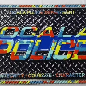 Puede incluir: Placa metálica rectangular con las palabras "OCALA POLICE" en letras coloridas y texturizadas. El fondo es negro con un patrón de placa de diamante. También se ve el texto "OCALA POLICE DEPARTMENT" e "INTEGRITY * COURAGE * CHARACTER".