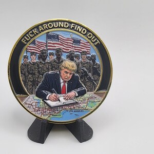 Puede incluir: Una moneda redonda con borde dorado, que representa a un hombre firmando un documento, con soldados y banderas americanas de fondo. La moneda tiene la frase "FUCK AROUND FIND OUT" alrededor del borde.