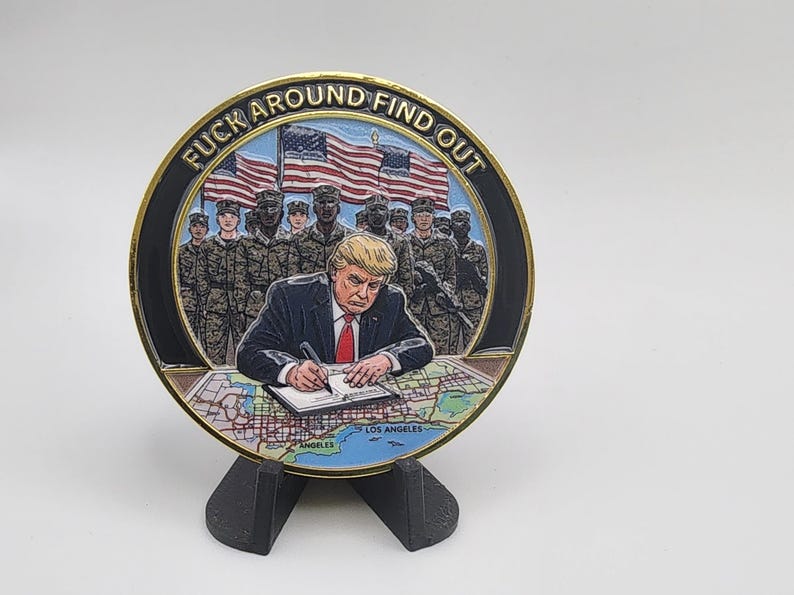 Puede incluir: Una moneda redonda con borde dorado, que representa a un hombre firmando un documento, con soldados y banderas americanas de fondo. La moneda tiene la frase "FUCK AROUND FIND OUT" alrededor del borde.
