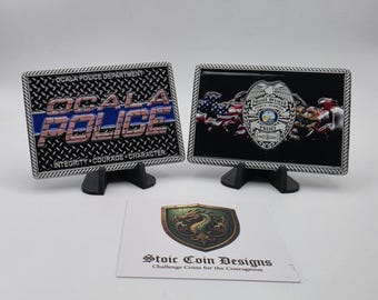 May include: Two rectangular metal plaques with silver borders, one with "OCALA POLICE" in brick-style letters and the other with a police badge and American flag imagery. The text "INTEGRITY COURAGE CHARACTER" is on the first plaque. A card with a dragon shield logo is in front.