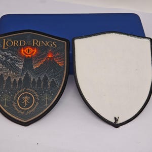 Puede incluir: Dos parches en forma de escudo con bordes negros. El parche izquierdo presenta una ilustración detallada de The Lord of the Rings con un ojo ardiente, una montaña oscura y un grupo de figuras. El parche derecho tiene un reverso blanco liso.