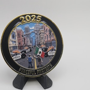 Puede incluir: Una moneda conmemorativa con borde dorado y borde negro, que representa una escena urbana con coches de polic&iacute;a y manifestantes. La moneda tiene el a&ntilde;o 2025 y la inscripci&oacute;n "Peaceful Protests". Se ve una bandera mexicana.