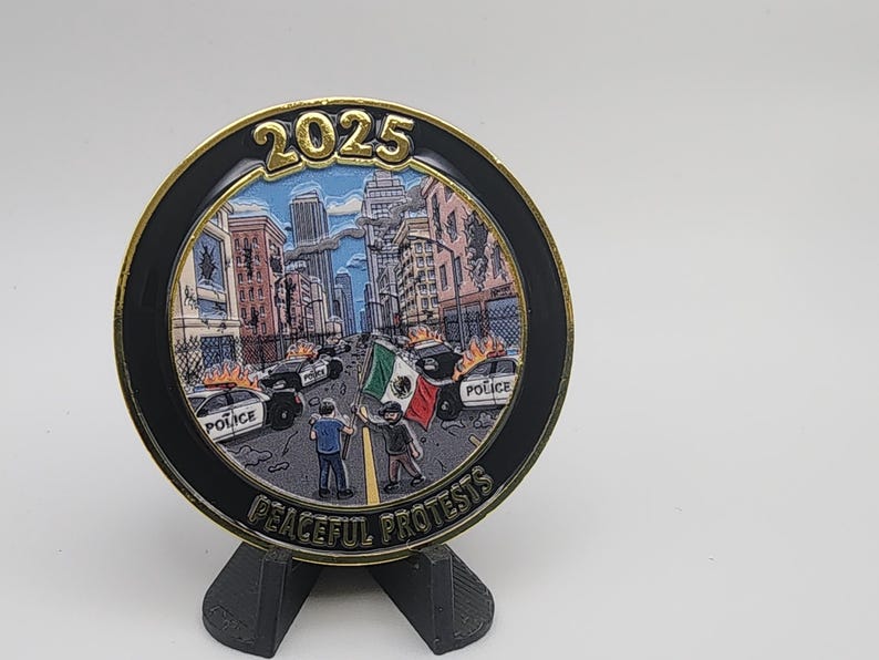 Puede incluir: Una moneda conmemorativa con borde dorado y borde negro, que representa una escena urbana con coches de polic&iacute;a y manifestantes. La moneda tiene el a&ntilde;o 2025 y la inscripci&oacute;n "Peaceful Protests". Se ve una bandera mexicana.