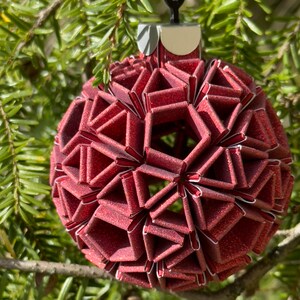 Pode incluir: Um enfeite de Natal vermelho e brilhante em origami com a forma de uma esfera. O enfeite é feito de papel dobrado e tem um design geométrico.