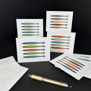 May include: A collection of white square greeting cards with a colorful abstract design of elongated shapes in blue, green, orange, and yellow. A gold pen and white envelopes are also present, all set against a black background.
