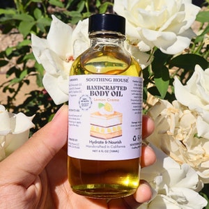 May include: A glass bottle of "Lemon Creme" body oil, held against a backdrop of white roses. The label says "Soothing House" and "Hydrate & Nourish." The bottle contains 118ml of oil.