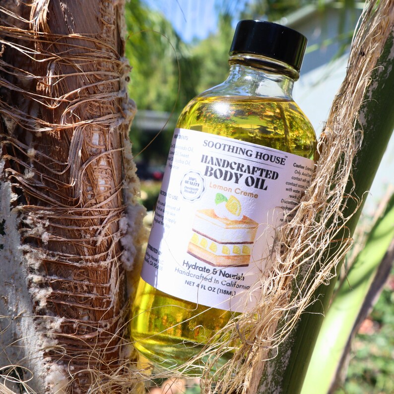 May include: A clear glass bottle of Soothing House Handcrafted Body Oil, Lemon Creme scent. The label features a lemon dessert illustration. The bottle contains 118ml of oil and is set against a natural background.