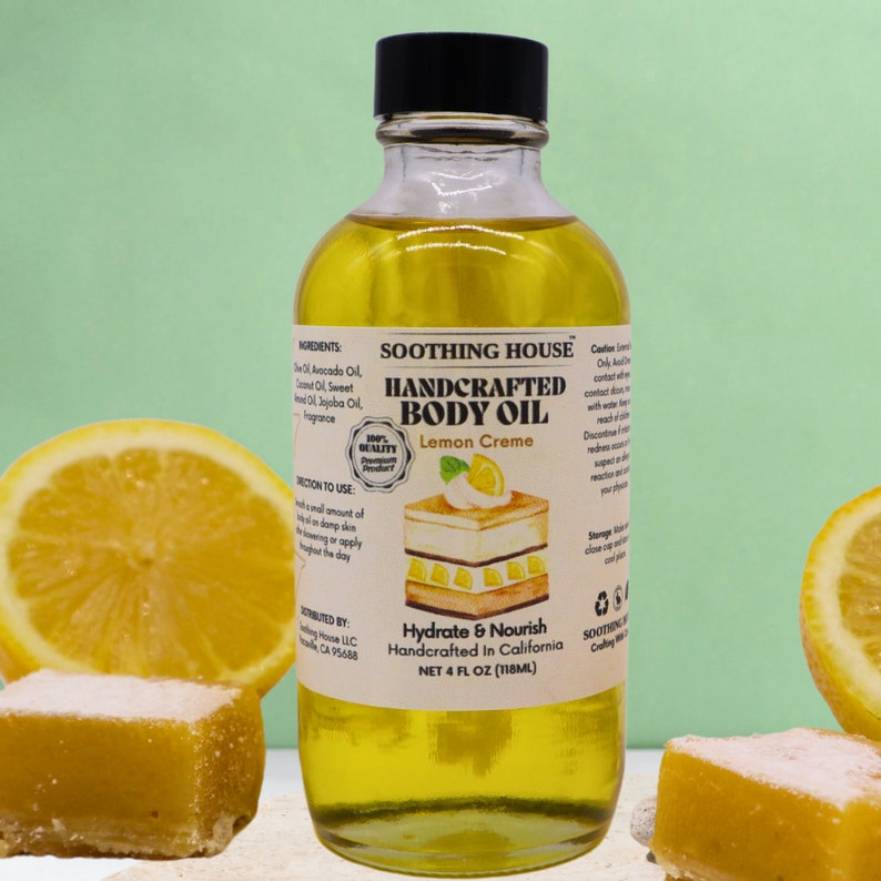 May include: A clear glass bottle of Soothing House Handcrafted Body Oil, Lemon Creme scent, with a black cap. The label includes the text "Hydrate & Nourish" and "NET 118ml". The bottle is surrounded by lemon slices and lemon-flavored treats.