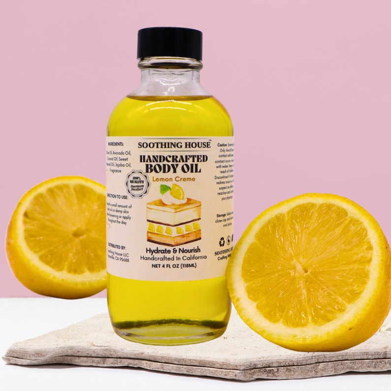 May include: A clear glass bottle of lemon creme handcrafted body oil with a black cap, next to two lemon halves. The label reads "SOOTHING HOUSE HANDCRAFTED BODY OIL Lemon Creme". The bottle contains 118ml of oil.