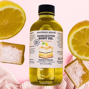 May include: A clear glass bottle of handcrafted body oil with a black cap, labeled "Lemon Creme." The label features a cake illustration and the words "Hydrate & Nourish." The image includes lemon halves and a bar of soap, all on a pink background. The bottle contains 118ml.