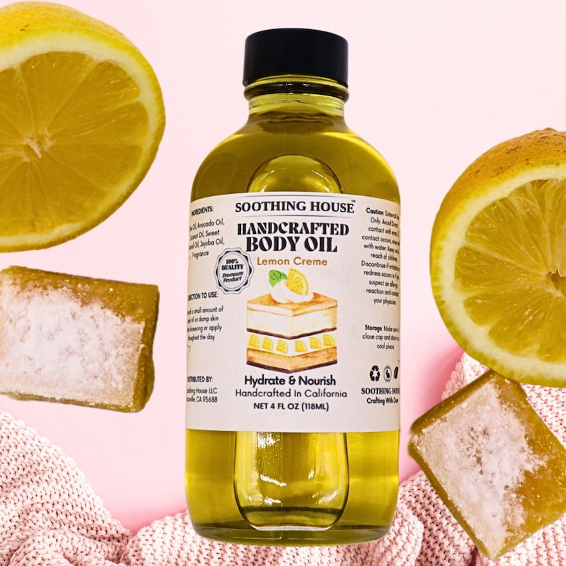 May include: A clear glass bottle of handcrafted body oil with a black cap, labeled "Lemon Creme." The label features a cake illustration and the words "Hydrate & Nourish." The image includes lemon halves and a bar of soap, all on a pink background. The bottle contains 118ml.