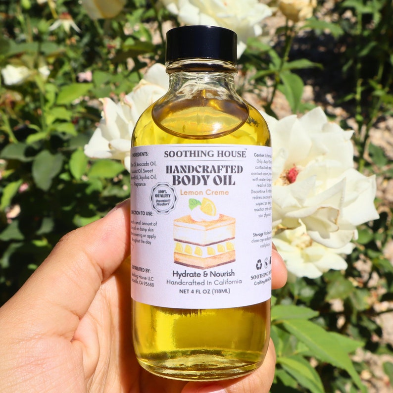 May include: A clear glass bottle of handcrafted body oil with a black cap, held in a hand. The label reads "Lemon Creme" and "Hydrate & Nourish." The bottle contains 118ml of yellow oil. The background features white roses.