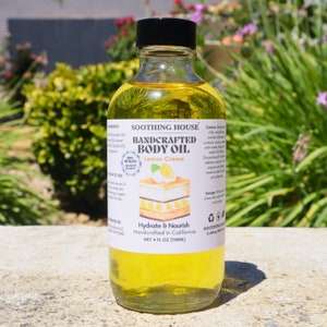 May include: A clear glass bottle of handcrafted body oil with a black cap. The label reads "Lemon Creme" and "Hydrate & Nourish." The bottle contains 118ml of yellow oil. The background features green foliage and sunlight.
