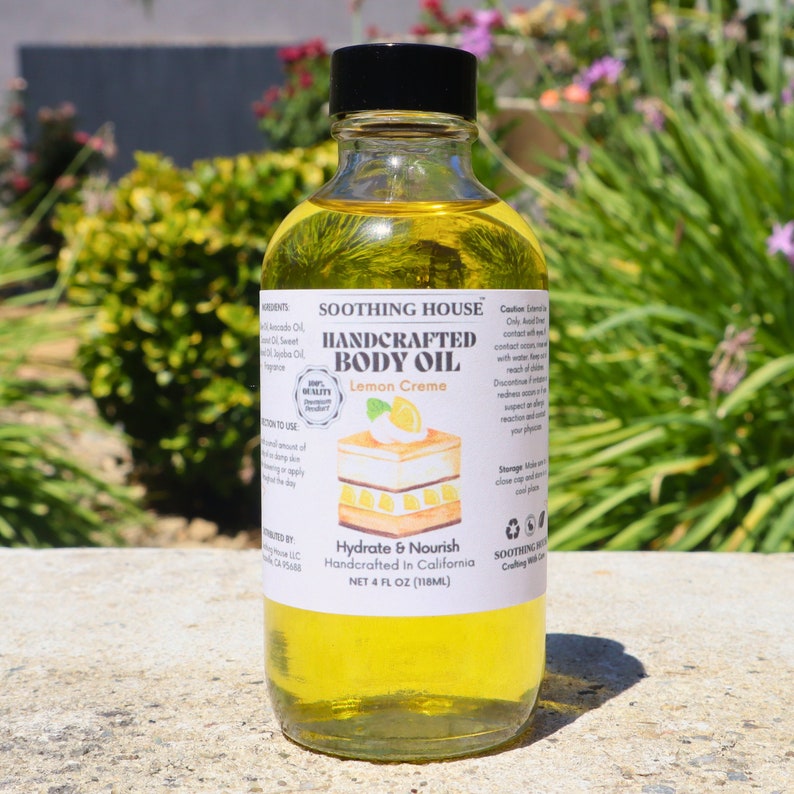 May include: A clear glass bottle of handcrafted body oil with a black cap. The label reads "Lemon Creme" and "Hydrate & Nourish." The bottle contains 118ml of yellow oil. The background features green foliage and sunlight.