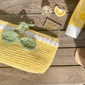 K&ouml;nnte beinhalten: Eine gelbe, geh&auml;kelte Tasche in Zitronenform mit gr&uuml;nen Bl&auml;ttern und wei&szlig;em Besatz. Die Tasche liegt auf einer Holzoberfl&auml;che mit einer kleinen Flasche Sonnencreme, einem Kamm und einer kleinen Zitronen-Dekoration.