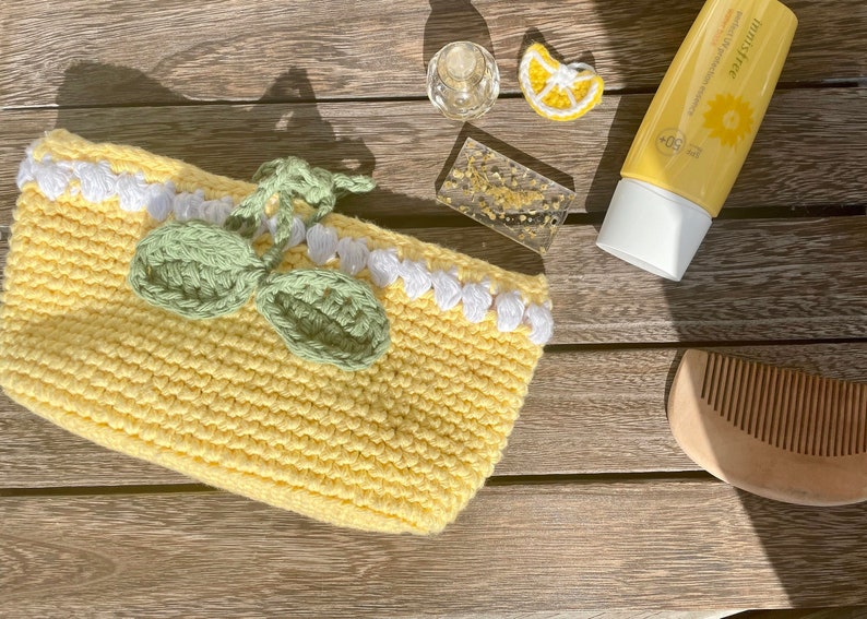 K&ouml;nnte beinhalten: Eine gelbe, geh&auml;kelte Tasche in Zitronenform mit gr&uuml;nen Bl&auml;ttern und wei&szlig;em Besatz. Die Tasche liegt auf einer Holzoberfl&auml;che mit einer kleinen Flasche Sonnencreme, einem Kamm und einer kleinen Zitronen-Dekoration.