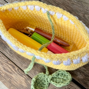K&ouml;nnte beinhalten: Eine gelbe H&auml;keltasche mit wei&szlig;en H&auml;kelbl&uuml;ten am oberen Rand. Die Tasche ist gef&uuml;llt mit verschiedenen Make-up-Artikeln, darunter Lippenstifte und Lipgloss.