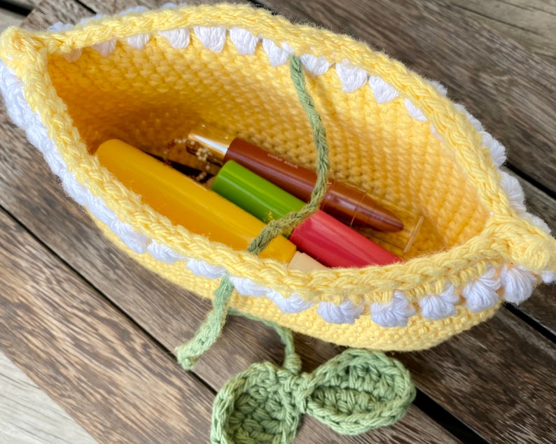 K&ouml;nnte beinhalten: Eine gelbe H&auml;keltasche mit wei&szlig;en H&auml;kelbl&uuml;ten am oberen Rand. Die Tasche ist gef&uuml;llt mit verschiedenen Make-up-Artikeln, darunter Lippenstifte und Lipgloss.