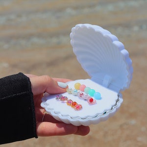 May include: A white seashell-shaped jewelry box with a variety of colorful earrings and pearl beads inside. The box is open and the earrings are arranged in rows.