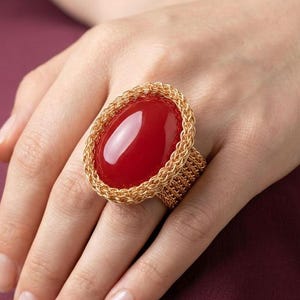 Könnte beinhalten: Ein Ring mit einem großen, glatten, ovalen roten Stein, der in einen strukturierten, goldfarbenen Rahmen aus geflochtenem Metall eingefasst ist. Die Ringschiene besteht ebenfalls aus demselben filigranen, goldfarbenen Drahtgeflecht.