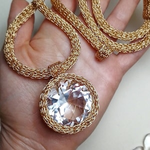Puede incluir: Un collar de cadena dorada con un gran colgante de cristal transparente. La cadena está hecha de pequeños anillos unidos y el colgante está engastado en un marco dorado.