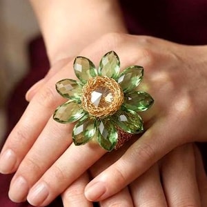 Puede incluir: Un anillo llamativo con un diseño en forma de flor, compuesto por pétalos de cristal verde translúcido y facetado que rodean un nido central de alambre dorado texturizado, con un cristal transparente de talla redonda en el centro.