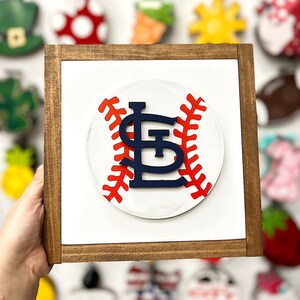 May include: A wooden framed sign with a white background featuring a red and white baseball with a blue "St. Louis" logo.