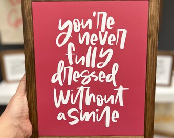 May include: A wooden framed sign with a pink background and white lettering that reads "you're never fully dressed without a smile".