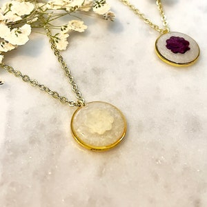May include: Two gold necklaces with round pendants. One pendant features a white pressed flower, the other a purple pressed flower.