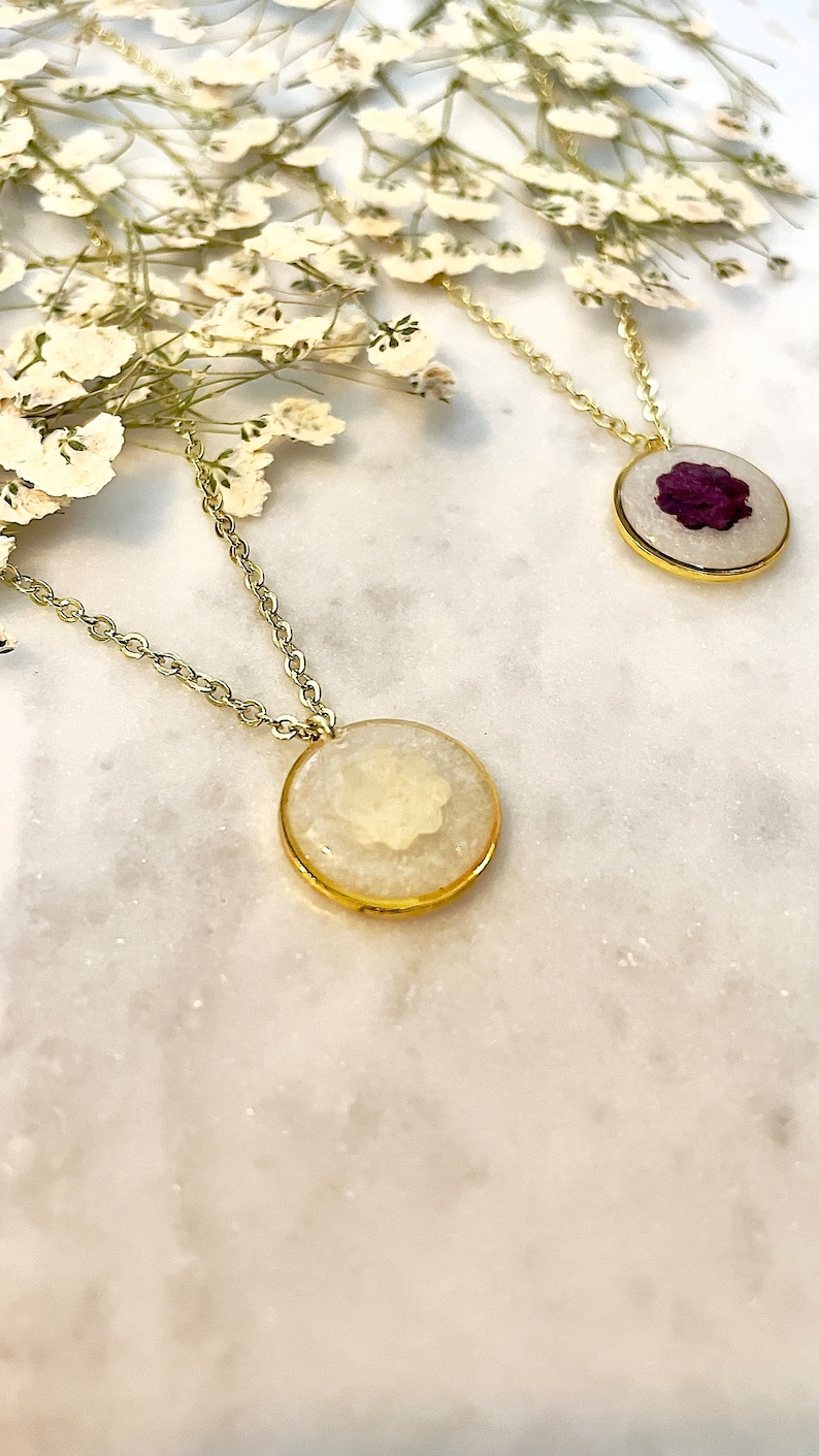 May include: Two gold necklaces with round pendants. One pendant features a white pressed flower, the other a purple pressed flower.