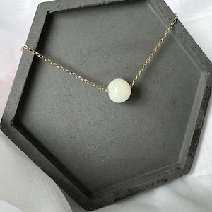 Pu&ograve; includere: Una collana a catena dorata con un'unica perla bianca perlacea. La collana &egrave; esposta su un vassoio esagonale grigio.