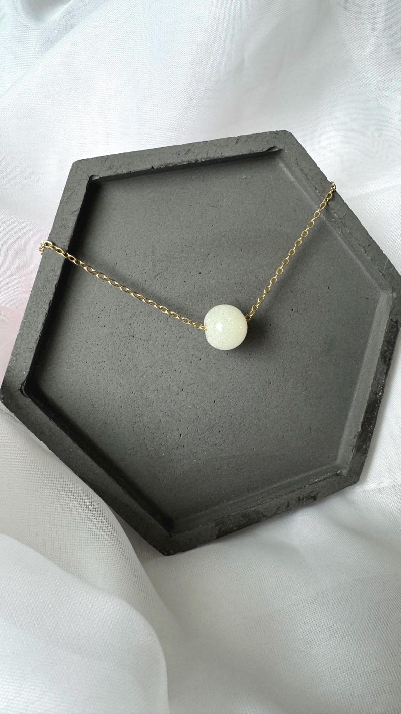 Pu&ograve; includere: Una collana a catena dorata con un'unica perla bianca perlacea. La collana &egrave; esposta su un vassoio esagonale grigio.