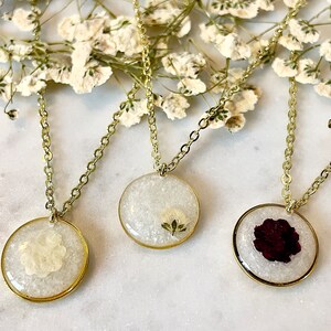 May include: Three gold chain necklaces with round pendants featuring pressed flowers. The pendants are filled with clear resin and contain a white flower, a green leaf, and a red rose.