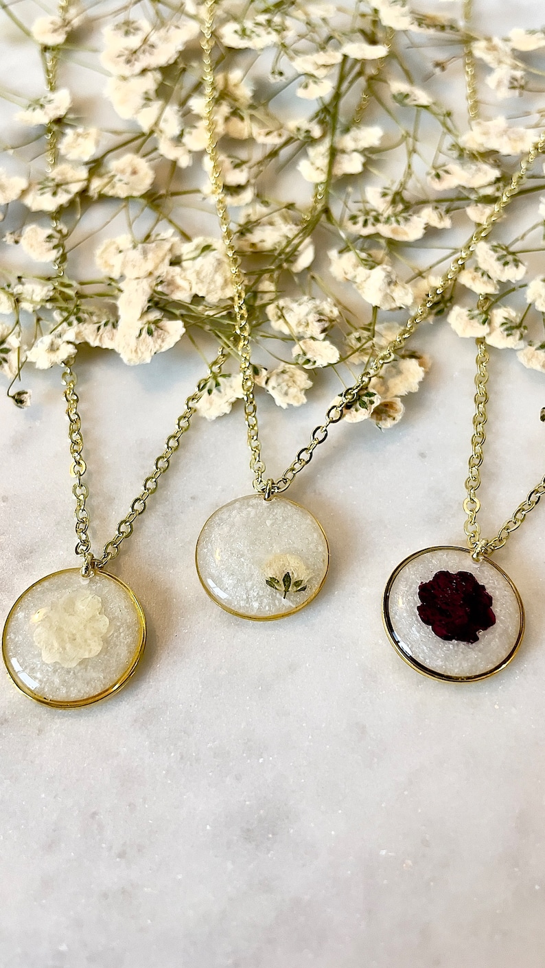 May include: Three gold chain necklaces with round pendants featuring pressed flowers. The pendants are filled with clear resin and contain a white flower, a green leaf, and a red rose.