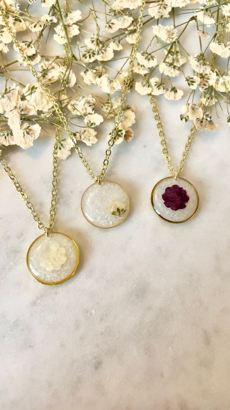 May include: Three gold necklaces with round pendants featuring pressed flowers. The pendants are filled with white glitter and contain a white flower, a white flower with green leaves, and a purple flower.