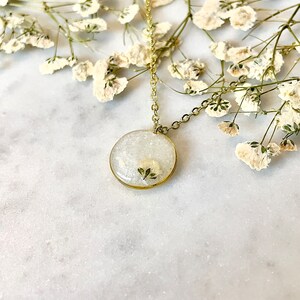 May include: A delicate gold chain necklace with a round pendant featuring a pressed white flower encased in clear resin.