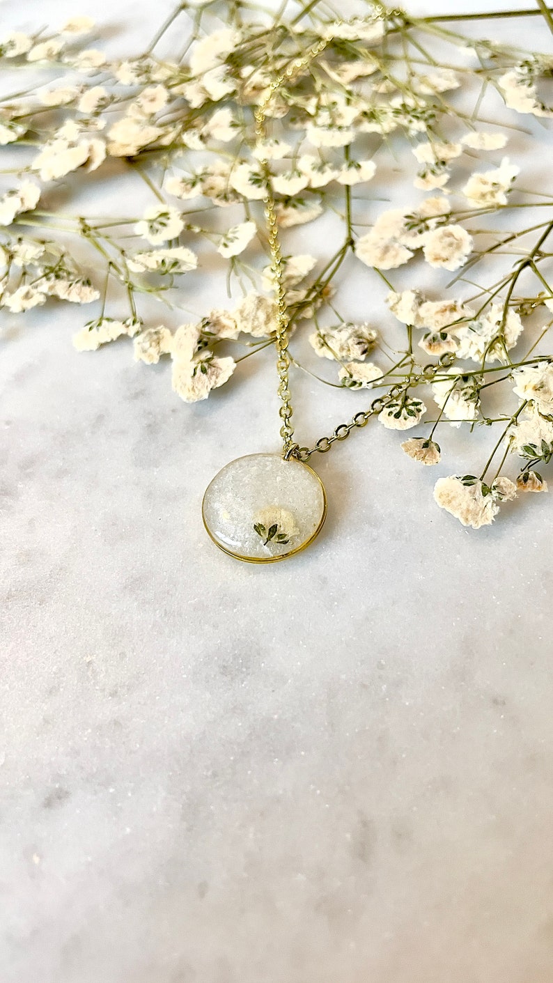 May include: A delicate gold chain necklace with a round pendant featuring a pressed white flower encased in clear resin.
