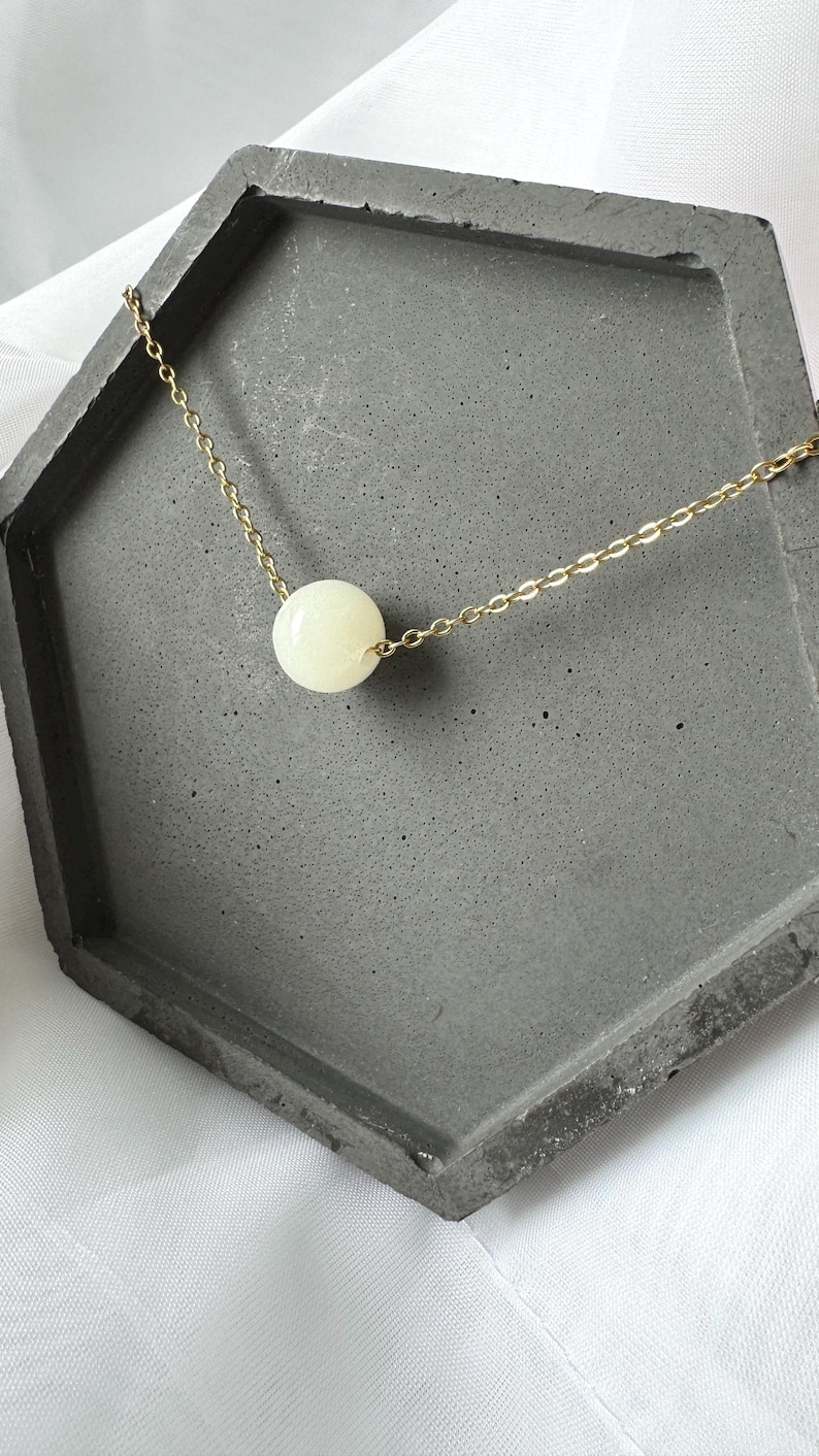 Pu&ograve; includere: Una collana con catena dorata con un unico grande ciondolo rotondo bianco.