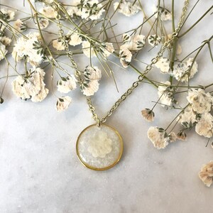May include: A gold chain necklace with a round pendant featuring a pressed white flower encased in clear resin.