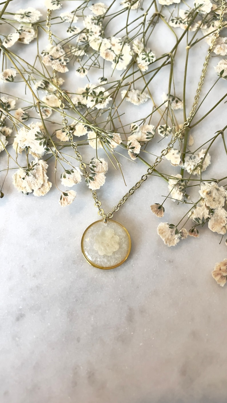 May include: A gold chain necklace with a round pendant featuring a pressed white flower encased in clear resin.