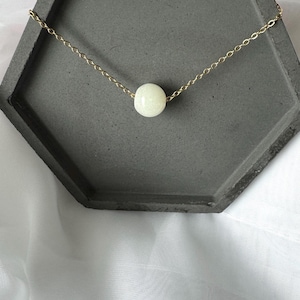 Pu&ograve; includere: Una collana a catena dorata con un solo ciondolo a forma di perla bianca. La collana &egrave; esposta su un vassoio esagonale grigio.