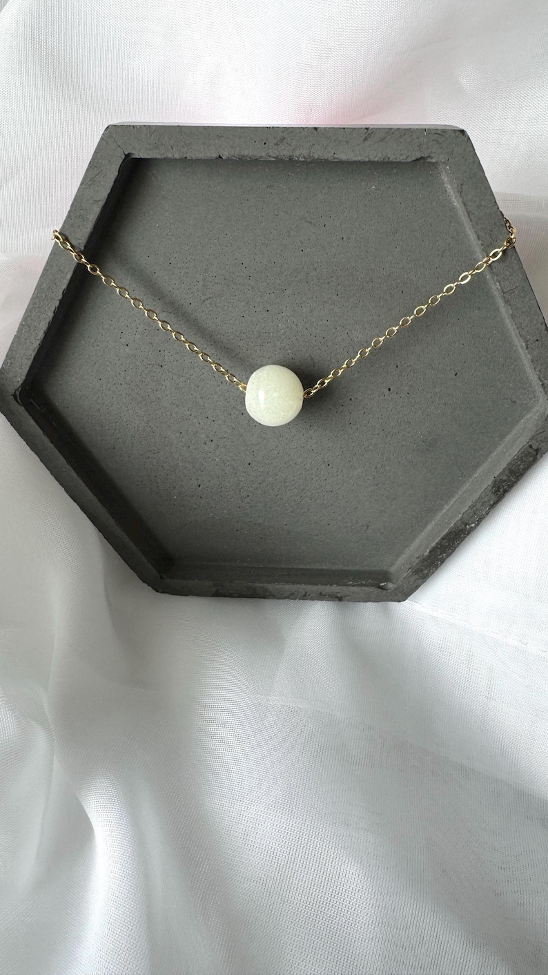 Pu&ograve; includere: Una collana a catena dorata con un solo ciondolo a forma di perla bianca. La collana &egrave; esposta su un vassoio esagonale grigio.