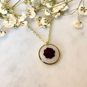 May include: A gold chain necklace with a round pendant featuring a pressed burgundy flower encased in clear resin.
