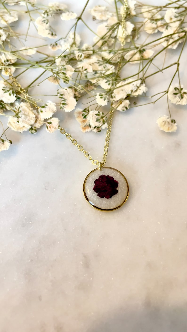 May include: A gold chain necklace with a round pendant featuring a pressed burgundy flower encased in clear resin.