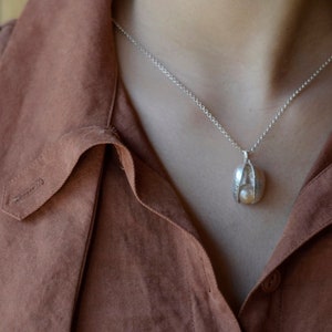 May include: A silver necklace with a unique, organic-shaped pendant. The pendant is made of silver and has a textured surface. The necklace is worn by a person wearing a brown shirt and blue overalls.