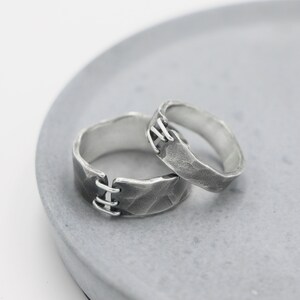 Puede incluir: Dos anillos de plata con un acabado texturizado. Los anillos están entrelazados y tienen un diseño de costura en la banda.