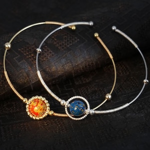 Op de afbeelding: Twee gouden armbanden met elk een klein, rond, kleurrijk sieraad. De sieraden zijn een blauwe en gouden bol en een rode en gouden bol.