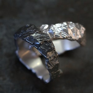 May include: Two textured metal rings, one dark gray and one silver, are intertwined. The rings have a rugged, organic appearance, with a rough, uneven surface. The interior of each ring is smooth and polished, creating a contrast with the exterior.