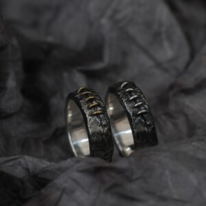 Puede incluir: Dos anillos de plata con un acabado texturizado. Un anillo tiene detalles dorados a lo largo de la costura.